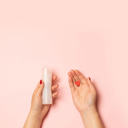 Womens hands hold intimate grease and a red heart shape on a pink background